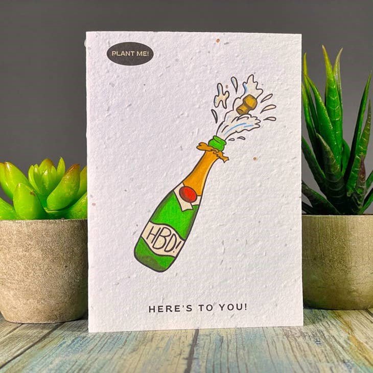 Seed paper birthday card featuring a green champagne bottle with HBD label and Here’s to you text. Wordkind.